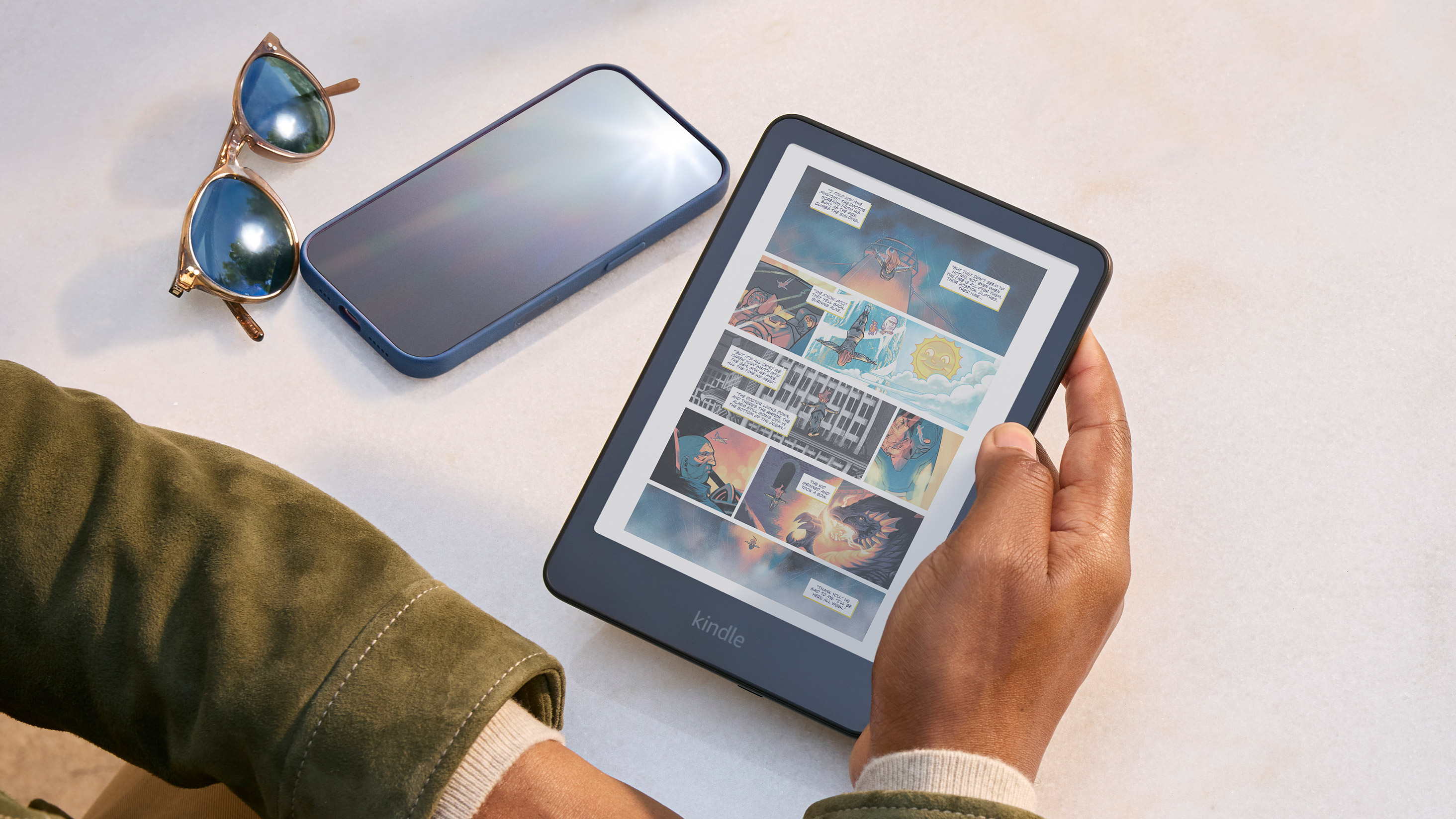 A reader holds Kindle Colorsoft while sitting at a table. A graphic novel is on screen. A phone and glasses are nearby, the screen not visible due to a glare.