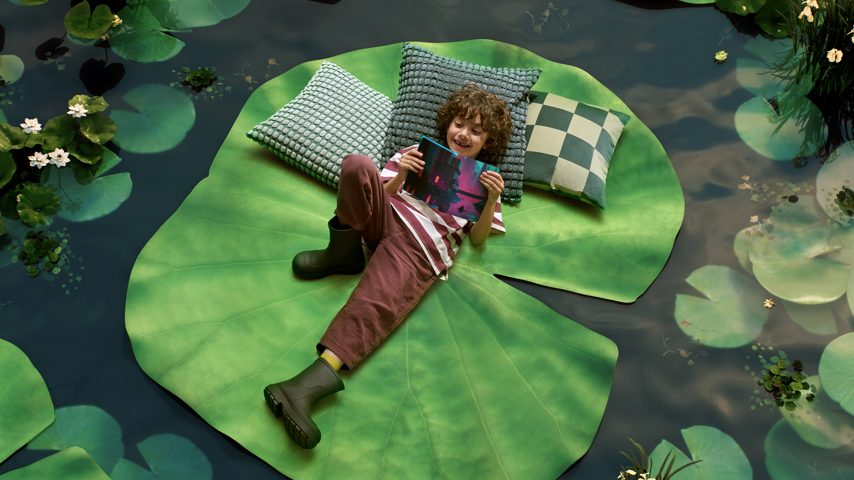Child lays on lily pad rug in middle of a pond floor and reads a Kindle Paperwhite Kids.