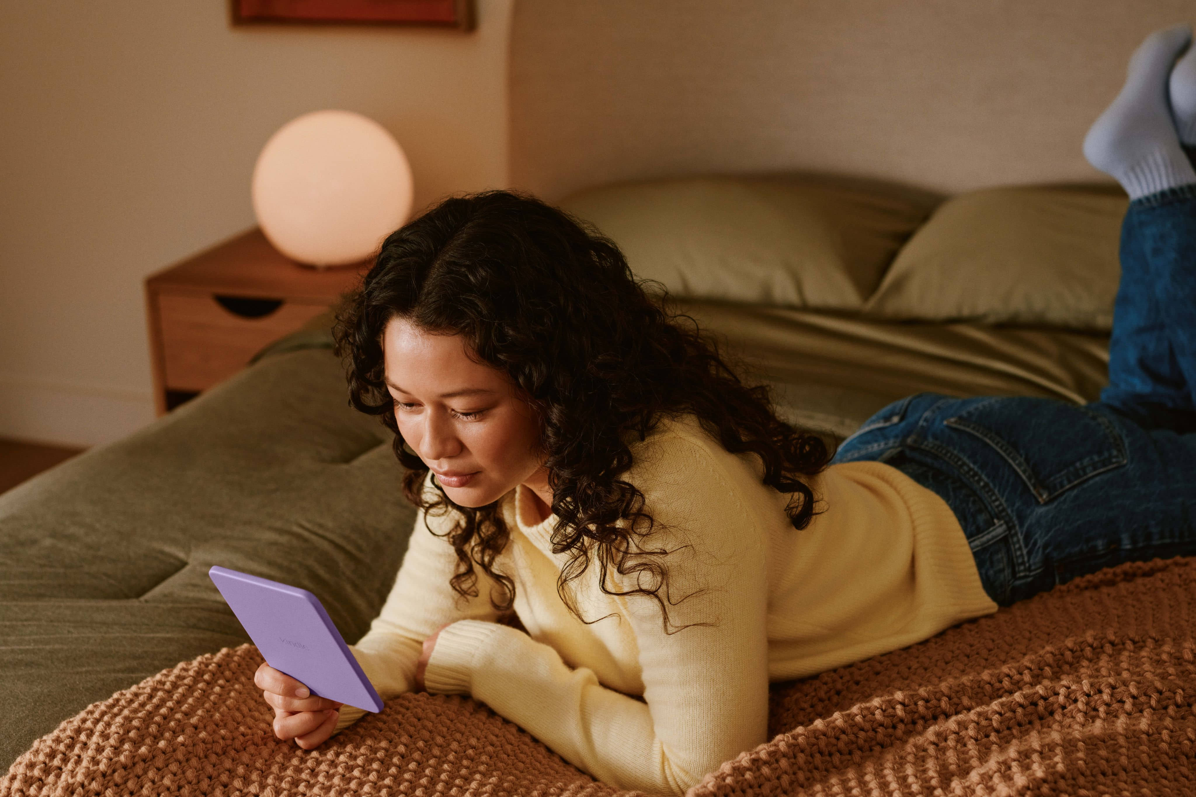 A person in a yellow shirt lying on a bed, reading on a purple Kindle.