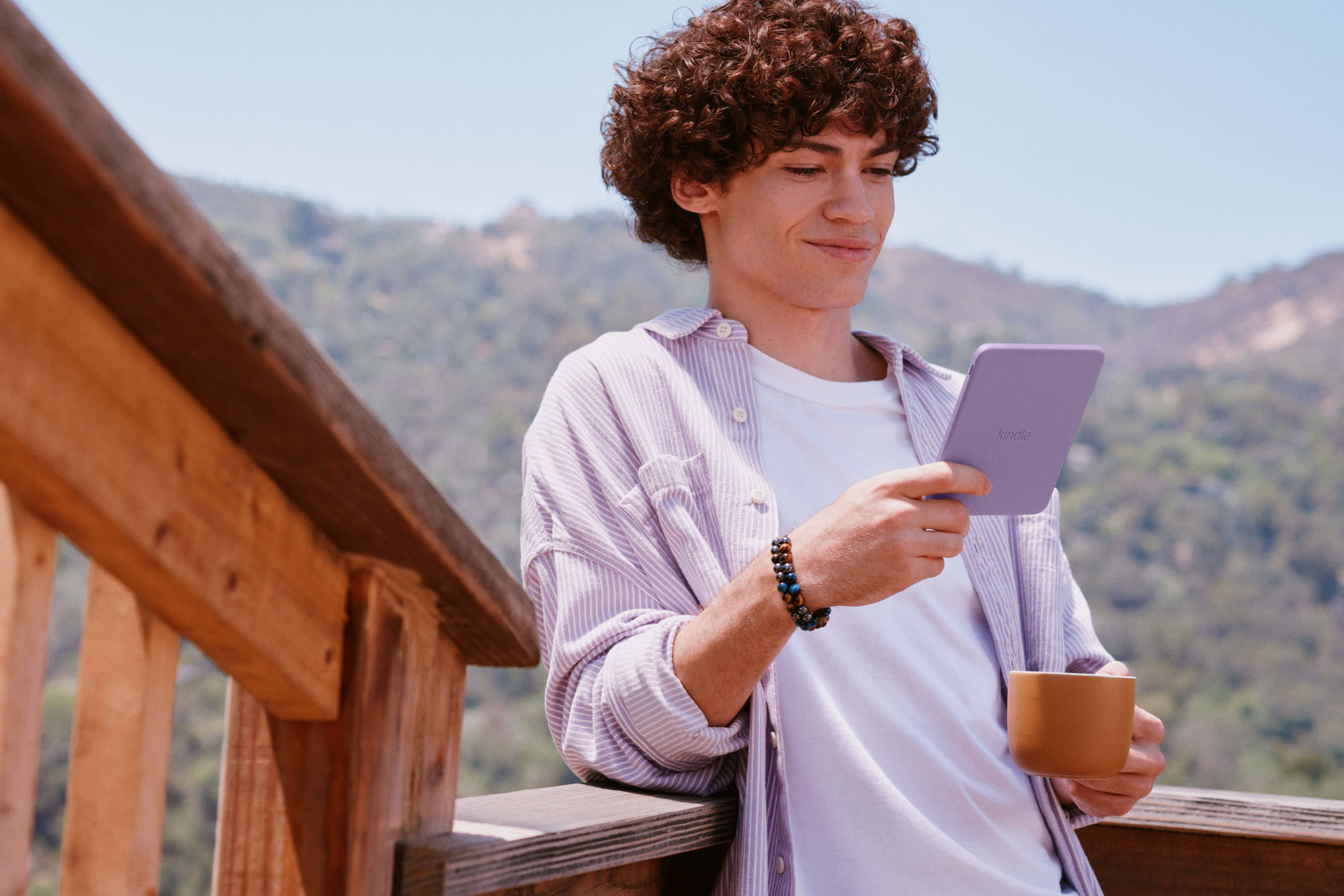 A person reading on a purple Kindle outdoors on a deck in bright daylight, mountains behind them.