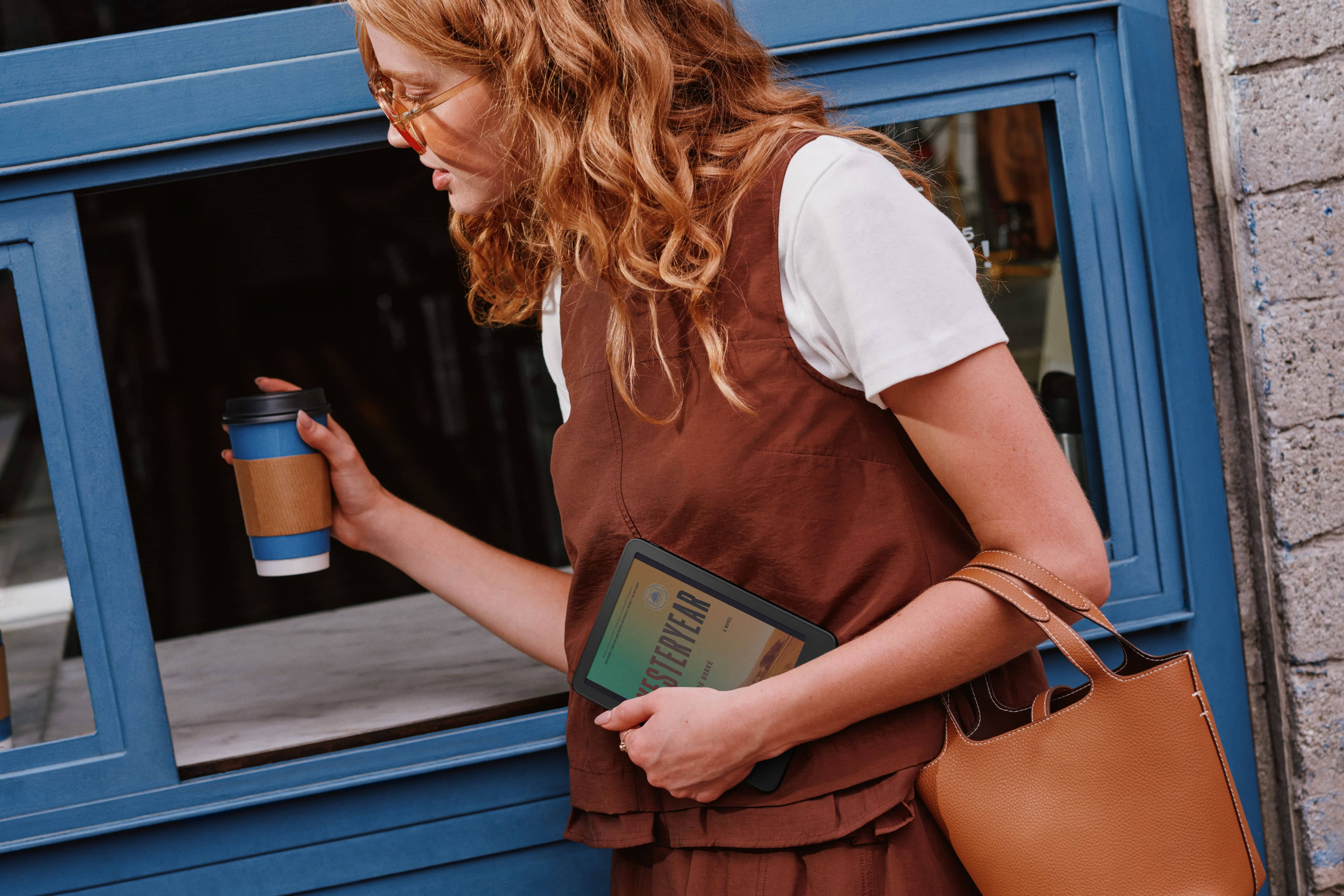 Person holding Amazon Kindle and coffee cup outdoors.