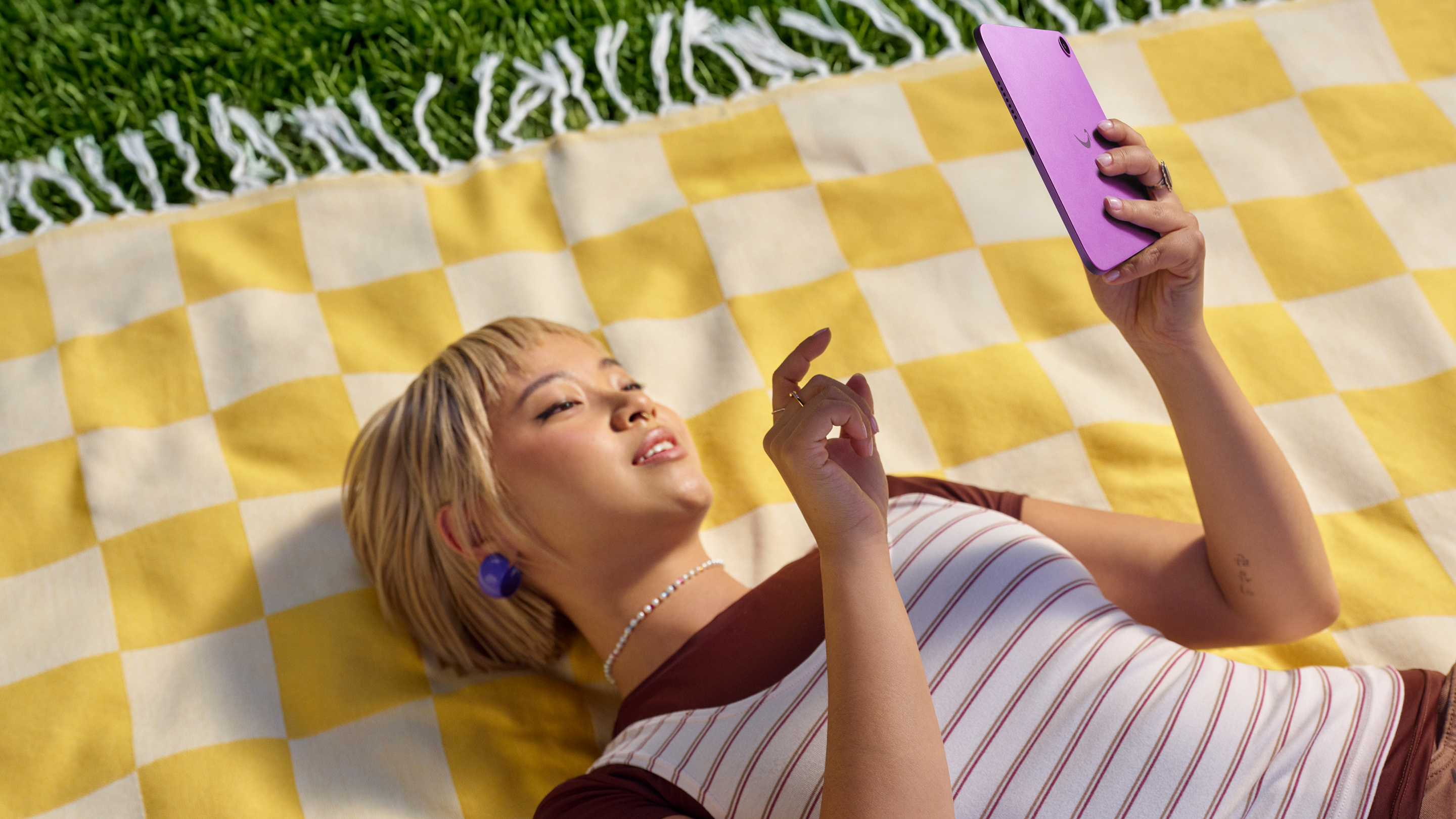 Outdoors, a person lays in a blanket on grass, holding an Amazon Alexa Tablet 8.